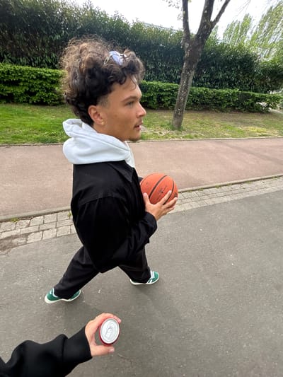 The image shows a first-person perspective, potentially a selfie, on a paved walkway in an urban area, specifically in Noisiel, France. The main subject is a young man with curly hair, an earring, and sunglasses resting on his head. He wears a white hoodie under a black jacket and black pants, coupled with green and white sneakers. He holds a basketball in one hand. Adjacent to him, another person is holding a red can, presumed to be a beverage. The background includes lush green foliage, trees, a sidewalk with a stone border, and a paved road. The overall scene suggests an outdoor recreational environment.