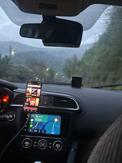 The image shows the interior of a car, likely taken from the driver's seat. The car is in motion, as the view through the windshield reveals a blurred landscape of green foliage and a road. It is raining, evident from the water droplets on the windshield.
The car's dashboard is visible, with a central infotainment screen displaying a navigation system showing a route approximately 31km to "Ресторант Пещерата" (Restaurant "Pescherata"). The navigation indicates the destination is in Lukovo, Bulgaria, arriving around 21:12, in 36 minutes. To the right of the screen are climate control buttons, set to 20 degrees Celsius.
A smartphone is mounted to the dashboard, displaying the Spotify application with various podcast covers. The steering wheel is partially visible at the bottom, and a rearview mirror is present in the upper-left corner of the frame.