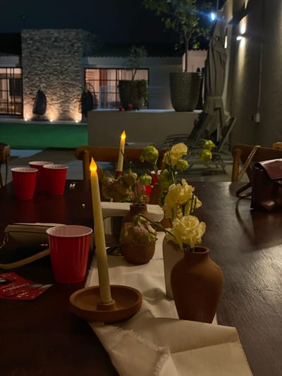 The image shows a table setting, likely outdoors, with decorative elements. There are clay vases, some with white flowers, and lit taper candles in wooden holders. Several red plastic cups are arranged nearby. A white cloth is draped on the table. In the background, a stone wall is visible, along with a building and some vegetation. The location appears to be Santa Cruz Tehuixpango, Mexico.