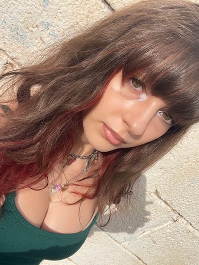A fair-skinned woman with brown hair and red highlights poses outdoors in a green ribbed tank top. She wears multiple necklaces with pendants, including a Star of David. Her makeup includes glossy lips and a light shimmer on her eyelids. Light reflects strongly off her left eye socket. The background features a textured, light-colored wall.