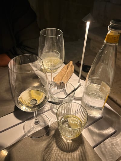The image shows a high-angle shot of a table setting, possibly in a restaurant, with elements suggesting it could be in Polignano a Mare, Italy.
There is an empty glass bottle with a yellow label. The label has the word “EL” on it, sitting sideways on the table. The bottle is clear.
There are three stemmed glasses on the table containing liquid, presumably wine. Some of the glasses are full and others are half full. There are also some empty glasses on the table.
There is a spoon resting in one of the glasses.
A folded brown napkin is visible among the glasses.
The table surface is white, and there are distinct shadows cast by a light source from above. The table appears to be made of wood or a composite material.
The backdrop seems to have a rough surface texture, possibly a stone wall, which is commonly found in Polignano a Mare.
The lighting is somewhat dim and warm.
The overall scene suggests a meal or gathering in a restaurant setting.