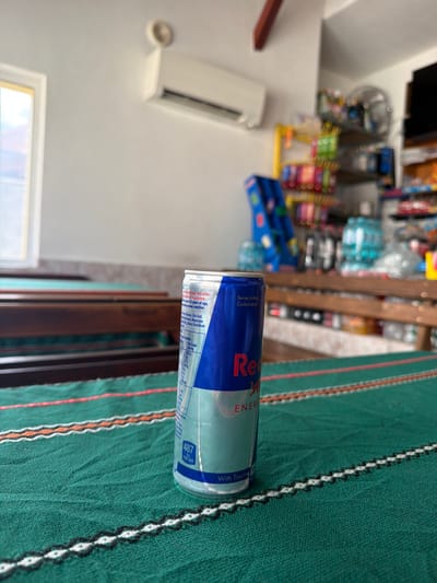 The image, set in Gradezhnitsa, Bulgaria, depicts an indoor commercial or recreational space. In the foreground, a silver and blue Red Bull energy drink can stands upright on a dark green fabric tablecloth. The tablecloth features horizontal stripes in orange, white, and black with intricate patterns. The can prominently displays "Red Bull ENERGY DRINK" text, a bull logo, and nutritional information, including "487 kJ per can" and "With Taurine." In the midground, dark wooden tabletops or benches extend horizontally across the frame. The background features a light-colored wall with a rectangular window on the left, partially obscured by bright sunlight. Above the window, a white wall-mounted air conditioning unit is visible. To the right, several shelves are stocked with various packaged consumer goods, including snacks and multiple large bottles of water. A yellow display stand and a black electric fan are also discernible amongst the merchandise. No individuals are present in the scene.