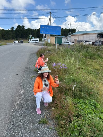 In T/B Seminskii Pereval, Russia, two women are captured in a roadside scene. The woman in the foreground, wearing an orange jacket, a light-colored hat, white pants, and pink sneakers, is squatting near a patch of overgrown grass and wildflowers. She is holding a disposable coffee cup and her phone, which is angled towards the flowers. Behind her, a second woman, also in an orange jacket and jeans, squats closer to the road, holding her phone up as if taking a picture. The road, composed of asphalt and gravel, curves to the left. A blue directional sign with Russian text indicates directions and distances. A cluster of trees forms a backdrop to the scene, with a few buildings and parked vehicles visible in the distance under a cloudy sky.