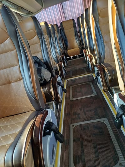 The image shows the interior of a bus in Bolshoy Sochi, Russia. The bus features rows of empty, brown and black leather seats with prominent stitching. The seats are designed with high backs and armrests. In the foreground, a seatbelt is visible. The floor is covered with a dark brown carpet with a subtle striped pattern. Along the aisle, there are yellow and white striped markings and metallic floor grates. The windows are covered with striped curtains. The lighting suggests it is daytime, with natural light filtering through the curtains, casting shadows on the seats. There are no people visible in the image.