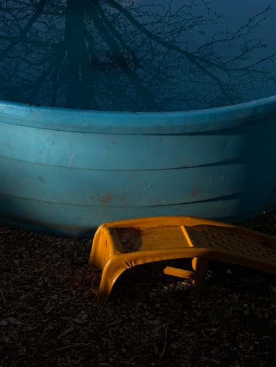 The image depicts a blue plastic kiddie pool resting on a dark ground surface with what appears to be wood mulch. Reflected in the blue plastic of the kiddie pool is the stark silhouette of bare tree branches against a deep blue sky, possibly captured at dusk or dawn. To the right of the kiddie pool, a section of a yellow plastic child's slide is visible, extending onto the mulch surface. The mulch has a mix of shades of brown, typical of decomposed wood chips. The scene is partially illuminated, creating distinct shadows that highlight the textures of the surfaces. The exact location within Boulder, United States, cannot be determined from the image alone. There are no people, subjects, actions, or interactions depicted in the image. The objects present are a kiddie pool, a section of a child's slide, and wood mulch. The environment is outdoors, likely a backyard or playground.