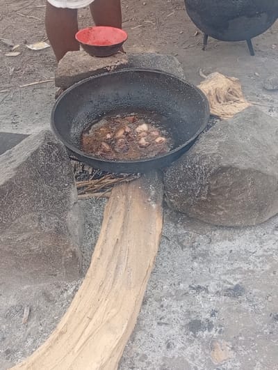 The scene is a cooking fire in the city of Assai Sabongida, Nigeria, located at coordinates (latitude: 8.864, longitude: 8.442). A large cast iron pot sits on top of three rocks. There is a wooden log underneath the pot and a red bowl is sitting next to the pot. The pot is filled with hot oil and what appears to be pieces of meat. The fire has died down and there are ashes and embers around the pot. The ground is uneven and dirt. There are pieces of twigs and sticks scattered throughout the scene. There are no people visible in the image.