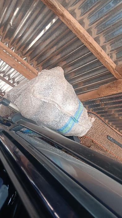 The scene, located in Langtang, Nigeria, depicts a large, off-white, woven polypropylene sack with prominent blue and green vertical stripes. The sack rests on a dark, reflective surface consistent with a vehicle's roof or a roof rack. Beneath the sack, a light-colored, cylindrical object and a brown, textured material are partially visible. Overhead, a corrugated metal roof, exhibiting slight undulations and dust accumulation, is supported by rough-hewn wooden beams. These beams show darker, linear patterns consistent with aged wood or environmental residue. The overall environment appears to be a sheltered or partially enclosed space, possibly an outdoor shed or carport. No individuals, overt actions, or interactions are present.