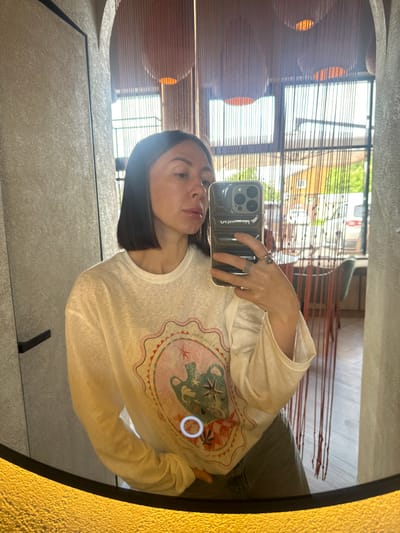 A medium shot captures a woman taking a mirror selfie in an indoor setting in Усады, Russia. The woman, with short, straight dark hair, is reflected looking towards the camera, holding a dark-cased smartphone (likely an iPhone) with her right hand, which displays multiple rings. She wears a white, slightly translucent, long-sleeved t-shirt featuring a colorful graphic design on the front depicting a framed image with an amphora and a star. Dark-colored pants are partially visible below her shirt.
The mirror's reflection reveals an interior with a textured light-colored wall and a dark vertical trim to the left, and an archway above. In the background, beyond the woman, large windows are visible, partially obscured by a brown, vertical string-like curtain or decorative divider. Through the windows, an outdoor scene is reflected, showing green foliage, residential buildings, and a white van. Interior elements reflected include recessed circular lights and what appears to be dining furniture (chairs and tables) in the distance. The bottom edge of the mirror features a distinctive textured, golden-yellow border. A white circular highlight is visible on the graphic design of the woman's shirt.