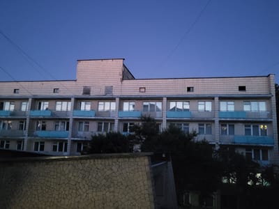 The image shows a multi-story building in the city of Khotsky, Ukraine, under a twilight sky. The building appears to be constructed with light-colored bricks and has multiple floors with numerous windows. Some of the windows have blue trim. Trees are visible in front of and around the building, partially obscuring the lower floors. In the foreground, there is a textured wall made of small, irregular stones. Power lines are strung across the sky above the building. No people are visible in the scene. The overall impression is one of an urban residential or institutional building in the evening.