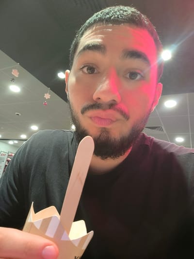 A man with a trimmed beard and short dark hair takes a selfie. He's holding a partly eaten ice cream with a wooden stick. The man is wearing a black t-shirt. The setting appears to be a brightly lit food court, with lights visible on the ceiling, and other people in the background. Red lighting casts an odd glow on his face. He's looking directly at the camera with slightly pursed lips.