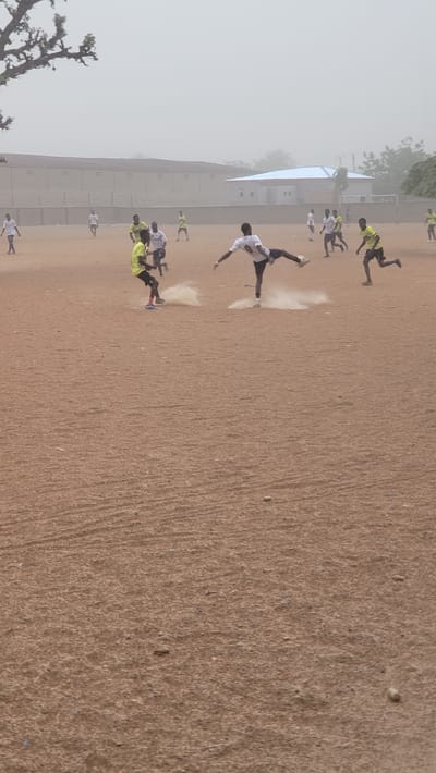 In Jogana, Nigeria, a soccer game unfolds on a dusty, brown field under a hazy sky. A group of players, some in yellow and black uniforms, others in white, are actively engaged in the game. A player in a white jersey with the number 8 is captured in mid-air, executing a bicycle kick, as dust rises from the ground. A building is visible in the background, partially obscured by the haze.