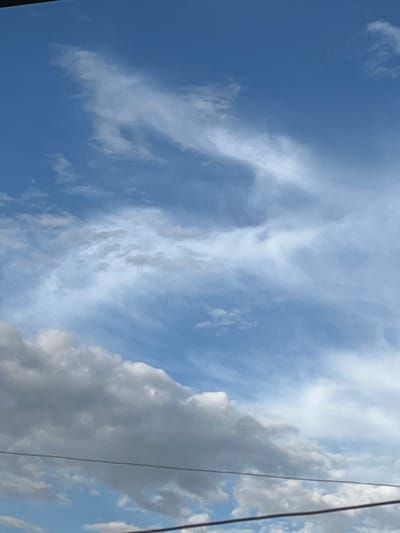The scene captures the sky above Jrarbi, Armenia. The upper two-thirds of the image show a clear blue sky interspersed with delicate, elongated white cirrus clouds. The lower third features denser, more substantial grey and white cumulus clouds. Two distinct, dark horizontal utility wires are visible spanning the lower portion of the frame. A thin, dark linear object is present along the extreme top edge. No human figures, animals, or specific activities are depicted.