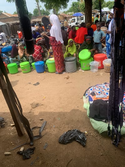 In Project, Nigeria, a bustling marketplace scene unfolds on a dusty dirt ground. The image captures a diverse group of individuals, some dressed in vibrant traditional clothing, others in more casual attire. A variety of large plastic containers, some green and others blue, are prominently displayed, suggesting they are for sale or transport. Amongst them, a woman in a red and white patterned dress and a white hijab is seen holding what appears to be a chicken. Nearby, a person in a green headscarf and dress is interacting with one of the green containers. In the background, a white truck is visible, along with several motorcycles, indicating a lively commercial hub. The overall atmosphere appears active and crowded, with numerous people milling about and engaging in various activities. The ground is uneven and scattered with debris, including plastic bags and what looks like dried corn husks, further emphasizing the outdoor market setting.