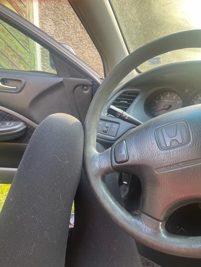 The image shows the interior of a Honda car. A person wearing black leggings is seated in the driver's seat. The Honda steering wheel is visible in the center of the frame, with the Honda logo prominently displayed. The dashboard and gauges are partially visible behind the steering wheel. Outside the car, one can see a blurry view of green grass and possibly a fence. The location in Blaskovo, Bulgaria is not discernible from the image content.