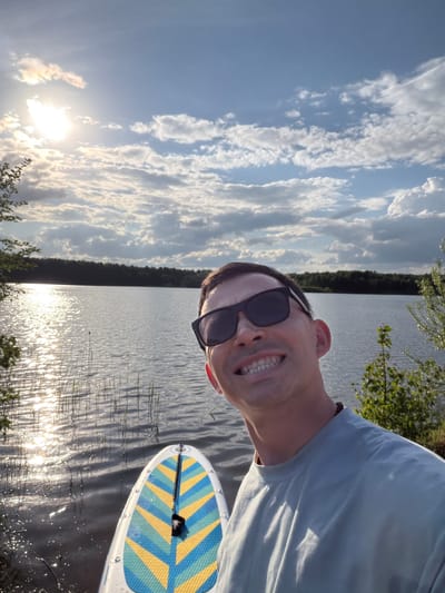 The image is a selfie taken on a lake, presumably in Selychka, Russia. A man wearing sunglasses and a light-colored t-shirt is visible in the foreground, smiling. A paddleboard is positioned adjacent to the man, partially visible in the lower right corner. The backdrop showcases a body of water with sunlight reflecting off its surface. The shoreline is lined with trees, and the sky is filled with numerous clouds. The sun is positioned low in the sky.