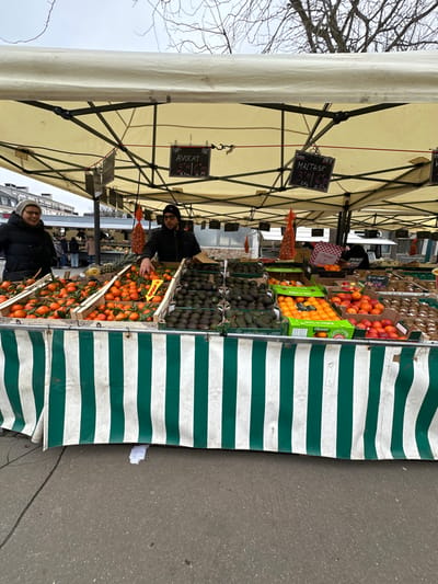 An outdoor market stall in Serris, France, is depicted under an off-white tent canopy. Two people are present at the stall. On the left, a woman wearing glasses and a dark winter coat stands, smiling towards the vendor. Behind the stall, a male vendor, wearing a dark beanie and jacket, is positioned with his right hand reaching into a crate of tangerines.

The stall's display features various fresh produce in wooden and cardboard crates. On the left and center, large quantities of tangerines with leaves are visible, alongside multiple crates filled with avocados. To the right, crates contain oranges, some explicitly labeled "Maltese," and other citrus fruits, with what appear to be kiwi fruits further right. Black chalkboard signs hang from the canopy, pricing "AVOCAT" (avocado) at 9€ per kilogram or 2€ per unit, and "MALTAISE" (Maltese orange) at 3€ per kilogram or 6€ per net. Red mesh bags, likely containing onions or similar produce, hang from the canopy near the center. The front of the market stall is adorned with a green and white vertical striped fabric skirt.

The ground in front of the stall is a dark paved surface, with a small piece of white litter visible. The background shows blurred urban buildings and bare tree branches against an overcast sky, consistent with an outdoor market environment.