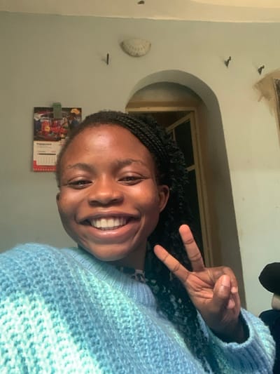 The image displays a young Black female situated indoors in Onibudo, Nigeria. She is positioned centrally, smiling broadly with visible teeth, and her left hand is raised, forming a "peace" sign. Her hair is styled in dark braids, and she is wearing a knitted sweater featuring horizontal stripes of light blue and turquoise. The background reveals a light-colored wall. On the upper left, a calendar, marked "ENGAGEMENT 2023," hangs. A shell-shaped wall sconce and several small, dark, hook-like fixtures are mounted on the wall. To the upper right, an arched doorway leads into an adjacent area, through which a window frame with a dark screen or glass is visible.