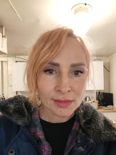A woman with short, peachy-blonde hair and light skin poses for a selfie in what appears to be a dimly lit interior space. She is wearing a dark blue jacket with a grey, furry collar and a pink and purple plaid lining. Her lips are painted a shade of rosy pink, and she has noticeable eyeliner and mascara on her eyes. The background suggests a functional or industrial setting, possibly a kitchen or workshop, with white cabinets, a bright overhead light, and some blurred objects on a counter. There are no visible signs of the city of Pavlokovo, Russia.