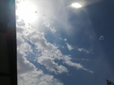 The image captures a daytime scene in Juma, Uzbekistan, dominated by a bright sky filled with scattered clouds. The sun is visible in the upper portion of the frame, partially obscured by clouds, creating a lens flare effect. The sky transitions from a lighter blue near the horizon to a deeper blue further up. On the left side of the image, a dark, angular structure, possibly part of a building or roofline, is partially visible, casting a shadow and framing the sky. The clouds are varied in texture and density, with some appearing wispy and others more cumulus-like. There are no people or animals present in the scene. The environment depicted is an outdoor urban or semi-urban setting.
