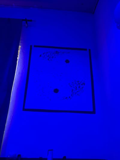 The image shows a dimly lit room bathed in a vibrant blue light, possibly from a blacklight or specialized LED lighting. The primary subject is a rectangular piece of artwork or a wall decoration mounted on the wall. This decoration is bordered by a thick black line and features two dark circular shapes, resembling eyes, set against a textured, dark background. The texture itself appears somewhat abstract and speckled, with small, irregular dark marks scattered across the surface.

To the left of the artwork, a portion of a door or cabinet is visible, painted a lighter shade that is also tinted blue by the ambient light. A small black object, possibly a hook or a latch, is attached to a surface below the artwork, on the left side of the frame.

To the right of the artwork, a white pipe or conduit runs vertically down the wall, partially obscured by the blue light.

The overall atmosphere is mysterious and somewhat abstract due to the intense blue lighting, which washes out any natural colors and creates strong shadows and highlights. The scene appears to be indoors, in a room with textured walls. The lighting suggests a deliberate setup, perhaps for artistic display or a specific mood. There is no visible text in the image. The location context of Pantinapu, Nigeria, does not provide specific visual cues within the image itself.