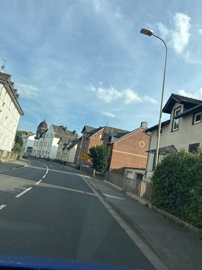 The image shows a street scene in Wetzlar, Germany, on a cloudy day. The perspective is from a vehicle looking out at the houses and road. The houses are of varying architectural styles, with some being older brick buildings and others more modern. A street lamp is visible in the upper left of the frame, casting a shadow over the street. The road is paved and has white lines marking lanes. There is some greenery visible, including trees and bushes, indicating a residential or mixed-use area. A yellow directional sign is visible on the right side of the street. The overall impression is of a quiet, everyday scene in a European town. There is no visible text in the image, aside from the potential text on the yellow sign, which is illegible. The time of day appears to be daytime, given the natural light and shadows.