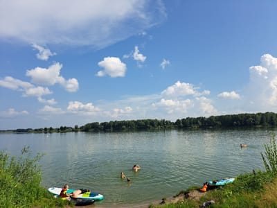 The image depicts a serene lake scene on a bright, partly cloudy day. In the foreground, on the grassy bank, are two paddleboards and a life vest. To the left, a person in a life vest is lying on a paddleboard in the shallow water, while another person lies on a separate paddleboard next to them. Further out in the lake, three individuals are swimming. In the distance, on the far side of the lake, a dense line of trees forms the horizon, with a few scattered buildings visible amongst them. A person is visible in the water on the right side of the lake, appearing to be using a floating ring. The lake water is calm with gentle ripples. The sky is a vibrant blue with scattered white cumulus clouds. There are no specific landmarks or identifiers to pinpoint the location within the city of Sorochii Log, Russia.