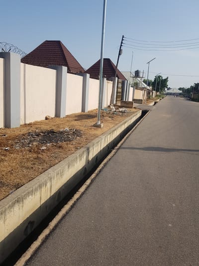 The image depicts a street scene in Palade, Nigeria, with a focus on infrastructure and the surrounding environment. A paved road runs diagonally through the frame, bordered by a concrete drainage channel on the left. Beyond the drainage channel is a low wall topped with barbed wire, with pyramidal roofs of buildings visible behind it. Litter and dry vegetation are scattered along the base of the wall. A lamppost stands near the foreground, and power lines stretch across the sky in the background. A few figures are walking in the distance on the road. The sky is clear and blue, suggesting a sunny day.