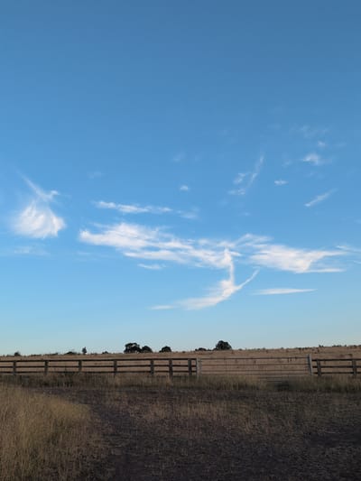 The image shows a rural landscape with a clear blue sky featuring wispy white clouds. In the foreground, there is dry, light brown grass and a dark, earthy path or barren ground. A wooden fence with metal gates runs horizontally across the middle of the frame, separating the foreground from a field of dry, golden grass dotted with a few trees on the horizon. The scene appears to be a tranquil countryside setting. The location is described as Little Wenlock, United Kingdom. There are no people or animals visible in the image.