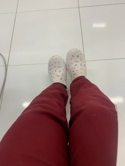 The image displays a first-person, downward-facing perspective of an individual's lower legs and feet within an indoor environment. The subject is wearing maroon trousers, consistent with medical scrubs, and white slip-on clogs. These clogs are decorated with a repeating pattern of small, multi-colored medical instruments, including syringes, stethoscopes, and pill bottles. The feet are positioned on a clean, reflective white tiled floor, segmented by thin dark grey grout lines. A slender grey cable lies on the tiled surface in the upper-left portion of the frame. The setting, suggested by the attire and footwear, indicates a medical or clinical facility. The specific location within Lauro De Freitas, Brazil, is not discernible from the visual information provided.