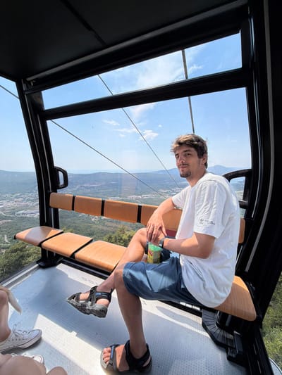 A man is seated inside a modern cable car, with large windows offering a panoramic view of a landscape below. The cable car appears to be in motion, indicated by the perspective of the landscape and the slight tilt of the image. The man is wearing a white t-shirt with an Adidas logo and some text, blue shorts, and sandals. He is holding a bottle of what appears to be a beverage. His gaze is directed towards the camera with a relaxed expression.
The scene inside the cable car shows a bench made of light-colored wood with black supports, and a textured floor. In the background, through the windows, a vast green valley dotted with buildings is visible, stretching towards distant hills under a clear blue sky with scattered white clouds. The overall impression is one of a scenic journey on a bright day. The presence of other people's feet in sandals and white sneakers at the bottom right corner suggests that there are other passengers in the cable car. The provided location context places this scene in Čavori, Montenegro.