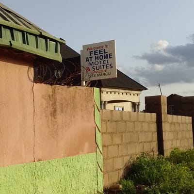 The image shows a white sign with blue and red text that reads "Welcome to: FEEL AT HOME MOTEL & SUITES OPPOSITE GSS MANGU." The sign is mounted on a wire fence, with barbed wire visible above it. The fence is attached to a stucco wall, painted a pale orange. To the right of the stucco wall is a wall made of concrete blocks, partially covered with greenery. In the background, a building with a dark roof and yellow walls is visible, with pillars supporting an overhang. The sky is a pale blue with some clouds. The scene appears to be in Mangu, Nigeria, as indicated on the sign.