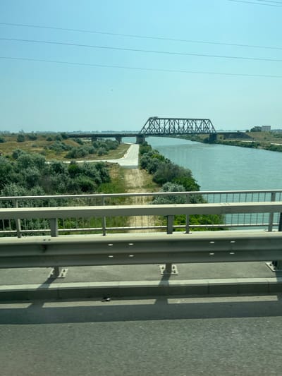 The image is a landscape view of a bridge over a river, likely taken from another bridge. In the foreground, a grey metal guardrail runs horizontally across the frame. Below the guardrail, a concrete barrier and a road surface are visible. The middle ground shows a wide river with shimmering water, bordered by lush green trees and vegetation on both banks. Two bridges span the river in the distance. The closest bridge is a dark metal truss bridge, and further back, a concrete bridge can be seen. The background features a hazy skyline with distant buildings and a clear, pale blue sky with thin power lines stretching across it. The scene suggests a sunny day in a semi-urban or rural environment, with a focus on infrastructure and natural elements. The presence of power lines and distant buildings indicates proximity to a populated area, and the overall setting is consistent with the area around Ovidiu, Romania, which is known for its bridges and proximity to water.