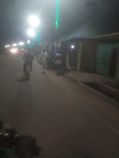 The image depicts a nighttime scene on a street in Tundun Wada West, Nigeria, illuminated by streetlights and the headlights of vehicles. Several individuals are visible on the street. In the foreground, a person is riding a motorcycle. Further down the street, a few people are walking, one of whom appears to be a man in a white garment. On the right side of the street, a building with a green door is visible. The overall environment suggests a moderately populated urban or semi-urban area at night.