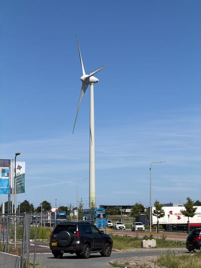 In Maasdijk, Netherlands, on a clear, sunny day with a blue sky, a tall white wind turbine stands prominently on the right side of the frame. Its three blades are extended, with the top one pointing upwards. A dark-colored SUV is parked on a paved road in the foreground, facing away from the camera, with its license plate clearly visible. Behind the SUV, several white cars are parked on the side of the road. In the mid-ground, a large white truck with red lettering that reads "HARP EXPEDITIE" is parked. A blue truck is also visible further to the left, along with other vehicles and buildings in the distance. Several tall trees are scattered throughout the scene. On the left side of the image, a row of flags are planted. The overall environment appears to be an industrial or commercial area, characterized by roads, vehicles, and large structures.