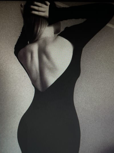The image is a grayscale, low-resolution photograph of a person's back. The person is wearing a black, long-sleeved dress with a deep V-shaped cutout that exposes their back and shoulders. Their hands are raised, with fingers interlaced at the nape of their neck, obscuring the top of their head. The dress accentuates the person's curves. The background is a textured wall. The image is of unclear origin and has no discernible connection to Chotěšov, Czechia.