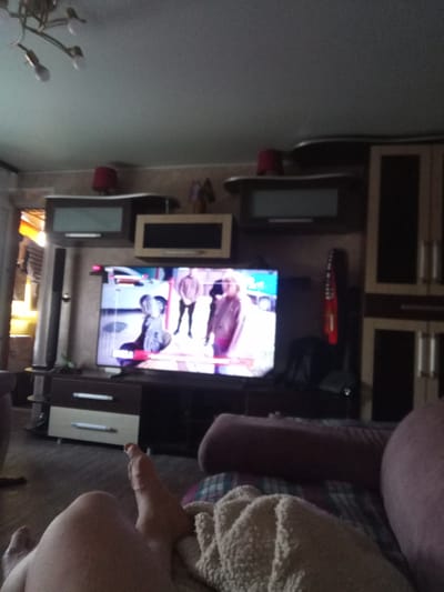 The image depicts a dimly lit living room in Mazyr, Belarus. The focal point is a large flat-screen television displaying a video with several people. To the right of the television is a tall, dark cabinet with multiple doors, possibly a wardrobe or storage unit. Above the TV, on a shelf, are two dark cabinets. Below the TV is a dark brown entertainment unit with two drawers.
In the foreground, a person's legs and feet are visible, suggesting they are lying down or relaxing on a couch or bed. The person is covered with a light-colored, textured blanket. The overall ambiance is casual and relaxed, with the television providing entertainment. The lighting in the room is low, creating a cozy atmosphere, and it appears to be either evening or a cloudy day. There are no visible signs of the weather outside. The style of the furniture and the general decor suggests a typical residential setting. No text is legible within the image.