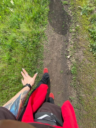 This is a first-person perspective image taken outdoors in Andreevka, Russia. The subject is walking on a dirt path, with lush green grass on the left and sparser vegetation and dirt on the right. The subject is wearing red shoes, black pants, and a red jacket or sweatshirt. Their left arm, adorned with a prominent blue and white tattoo, is visible, as is their left hand wearing a silver snake-shaped ring and a black smartwatch. The subject's right leg and foot are also visible, wearing a red shoe and black pants. The overall lighting suggests a sunny day.