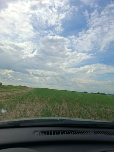 The image, likely taken from inside a vehicle, shows a view of the sky and a field. The sky is predominantly blue with a large number of white clouds. A field is seen on the right side of the frame; green plants are sparsely covering the brown earth. The right-most edge of the frame includes a portion of the car interior, specifically a gray panel with ventilation slots. The photograph seems to be taken at an angle, tilting to the left. Based on the prompt, the location is the city of Pobeda, Bulgaria.
