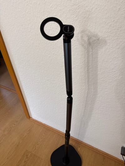 This media file shows a black articulating arm attached to a circular black base, which is resting on a wooden floor. The arm extends horizontally across the frame, reaching towards a white textured wall. The arm itself appears to be made of dark metal and has visible joints allowing for adjustability. On the left side of the arm, a circular attachment with the word "TELESIN" is visible. The lighting suggests an indoor environment, possibly during the daytime, as there are no distinct shadows indicating artificial lighting. The scene is static, with no people or active events depicted. The overall impression is a functional object, likely a mount or stand, positioned in a domestic or office setting. The floor has a wood grain pattern, and the wall has a textured finish.