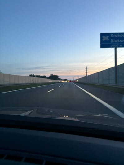 The image shows a view from inside a vehicle driving on a highway at dusk. A blue sign is visible on the right side of the road, indicating "Kraków" and "Biezanów" with distances of 3000 meters. The highway has multiple lanes and is bordered by sound barriers. The sky is clear blue, transitioning to a softer gradient of orange and pink near the horizon. Distant traffic lights and streetlights are visible in the distance. The foreground shows the interior of the vehicle, including the dashboard. The location is identified as Kokotów, Poland, along a highway leading towards Kraków.