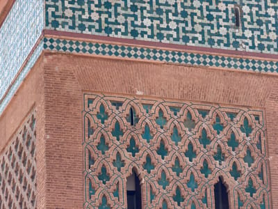 This close-up image showcases the intricate architectural details of a building located in Mechouar Kasbah, Morocco. The scene focuses on an exterior corner of the structure, revealing multiple layers of decorative elements.

The uppermost section features a wide band of geometric mosaic tilework, known as zelij, in vibrant shades of teal and off-white. This elaborate pattern forms interlocking stars and complex linear designs. Directly beneath this, a narrower horizontal border displays a repeating pattern of small, alternating teal and white diamond-shaped tiles.

Below these tiled sections, the building's wall is constructed from reddish-brown brickwork, showing variations in texture. An arched recess within this brickwork frames a large, intricately carved screen. This screen is composed of repeating lobed arches and stylized floral motifs, also in shades of reddish-brown with teal accents visible through the openings, suggesting a backing or integrated elements of contrasting color. This decorative screen likely serves as a window or ventilation grille. A small, vertical rectangular opening is also visible in the upper teal tilework.

The lighting is soft and diffused, indicating daytime conditions without harsh sunlight. There are no people or discernible activities taking place. No visible text is present. The distinct style of the tilework and brick patterns is characteristic of Moroccan Islamic architecture.