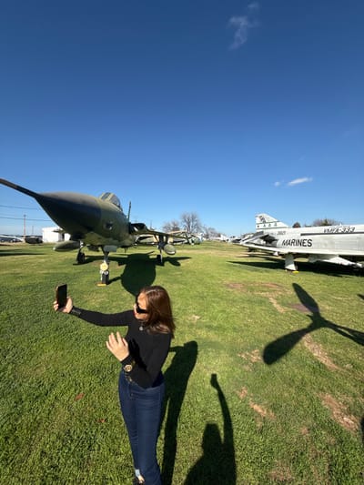 A woman in a black top and jeans photographs a military jet on a sunny day. The green and black jet, parked on a grassy field, faces toward the lower left corner. Behind the jet, other aircraft are visible. The sky is blue with a few scattered clouds. A shadow of the jet's propeller blades falls on the grass near a white plane labeled "Marines." The woman is in the lower right, crouching to take a photo with her phone.