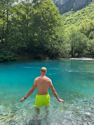 A shirtless man wearing neon yellow swim trunks stands in the crystal-clear, turquoise water of the Lake of the Gods in Vusanje, Montenegro. His back is to the camera as he looks out at the serene lake, with his arms extended outwards. The water's edge is lined with smooth, grey pebbles. Lush green trees cover the surrounding hillsides, which rise up to meet a rugged, mountainous backdrop under a bright, sunny sky.