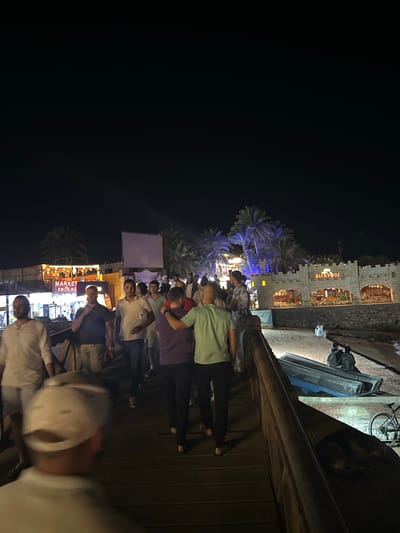 The image depicts a crowded outdoor scene in Dahab, Egypt, at night. A group of people stands on a wooden platform or bridge overlooking a street lined with shops and restaurants. The buildings are illuminated with bright lights and signs, including one that reads "Al Capone." Several palm trees are visible, some illuminated with blue lights. The sky is dark. Some of the individuals are looking down at the street, while others are engaged in conversation. A bicycle is visible on the bridge. The scene appears to be a tourist area.