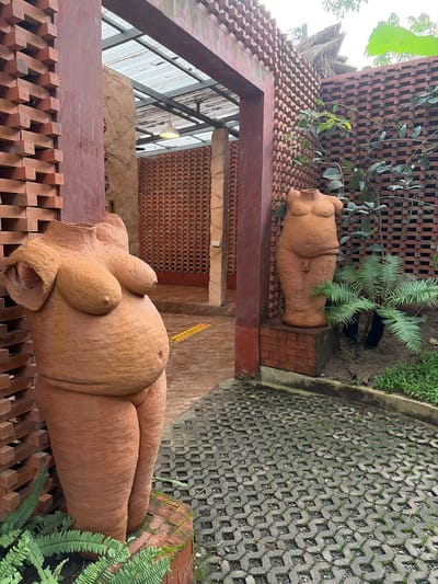 In Tambon Mae Raem, Thailand, a courtyard features two terracotta sculptures of the human torso and lower body. The sculpture in the foreground is a full torso, prominently displaying large breasts and a rounded belly. The sculpture in the background depicts the lower body and torso, including visible genitalia. The courtyard is paved with interlocking concrete blocks and surrounded by brick walls. Green plants, including ferns, grow in the garden beds around the sculptures. A covered walkway with a metal roof and fluorescent lighting is visible in the background. The overall impression is one of an outdoor sculpture garden or artistic installation.