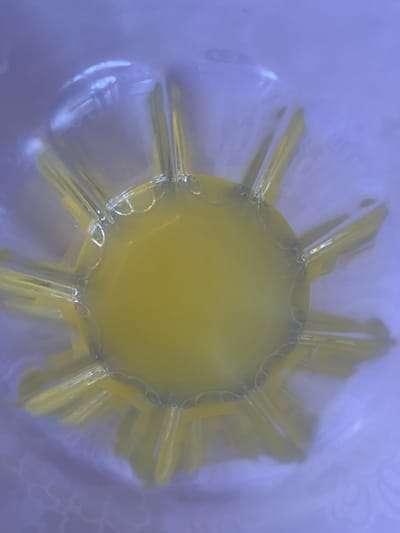 The image is an extreme close-up, top-down view of a bright yellow liquid, possibly egg yolk, contained within a clear, fluted vessel. The vessel has a starburst pattern of raised ridges radiating outwards from the center. Small bubbles are visible along the rim of the liquid where it meets the vessel's ridges. The background is a soft, out-of-focus lavender color, suggesting an ambient light source or colored background. The image lacks any discernible features to place it within a specific city or country.