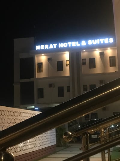 Illuminated vertical sign reads "MERAT HOTEL & SUITES" adjacent to a tall, white building with numerous windows and air conditioning units. A golden-colored metal railing runs diagonally across the right side of the frame, partially obscuring the view of the building. The setting appears to be nighttime or low-light conditions, emphasizing the brightness of the hotel sign. A patterned walkway is visible near the railing.