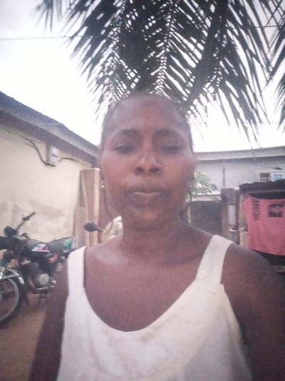 A woman is pictured in Kupai, Nigeria. She is wearing a white tank top and is standing outdoors. Behind her, there is a building and what appears to be a palm tree. A motorcycle is also visible in the background.