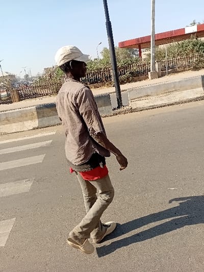 The image portrays a lone individual walking from left to right across a paved road under a clear sky in Sharada, Nigeria. The dark-skinned individual wears a light-colored baseball cap, a visibly worn muted brown or gray long-sleeved shirt with tears, light-colored trousers, and light-colored slip-on shoes, with the left shoe partially disengaged at the heel. A red undergarment is visible at the waistline. A light-colored object is present on the ring finger of the individual's right hand. The road surface features white zebra crossing markings on the left. A long shadow is cast on the road, indicating sunlight from the upper left. To the right of the road, concrete barriers separate the street from a raised area. Beyond these barriers, a dark metal fence borders dense green vegetation. In the background, a structure with a prominent red roof supported by white pillars, consistent with a fuel station canopy, is visible alongside utility poles.