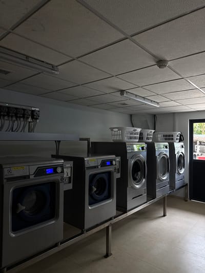 The image depicts a laundry room at an unknown location in the Maldives. The room contains several commercial-grade washing machines arranged in a row. Some of the machines are Electrolux Professional brand. White plastic laundry baskets are stacked on top of some of the machines. The room has a dropped ceiling with fluorescent lighting fixtures. There are also visible pipes and a dispensing unit on a shelf above the washing machines. A doorway is visible on the right side of the image, showing a glimpse of an outdoor area. The floor is tiled in a light color. The scene appears to be unoccupied.